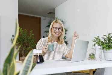 Happy freelancer with tea cup and laptop sitting at desk - TYF00575