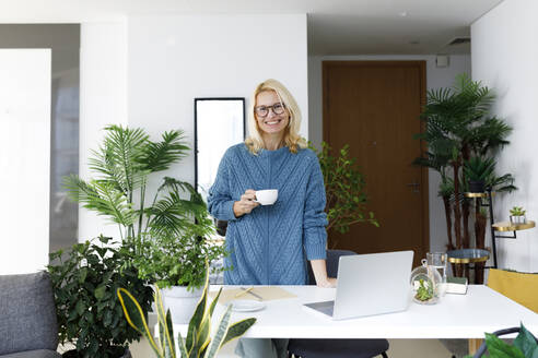 Glücklicher Freiberufler stehend mit Teetasse am Schreibtisch zu Hause - TYF00592