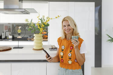 Smiling woman with smart phone and tea cup standing in kitchen - TYF00601