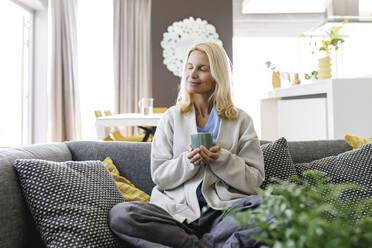 Smiling woman with tea cup sitting on sofa at home - TYF00611