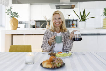 Lächelnde Frau mit Kaffee in Tasse und Kanne am Esstisch sitzend - TYF00614
