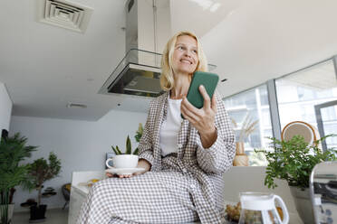 Smiling woman with smart phone and coffee cup sitting on kitchen counter - TYF00619
