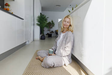 Smiling woman holding cup of tea and sitting with eyes closed on rug in kitchen - TYF00622