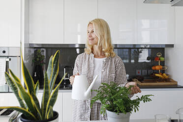 Lächelnde Frau mit Gießkanne, die neben einer Topfpflanze in der Küche zu Hause steht - TYF00623