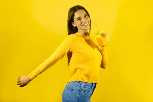 Glückliche junge Frau hält ein Glas Orangensaft vor gelbem Hintergrund - JSMF02617