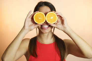 Playful young woman holding oranges over eyes against peach background - JSMF02618