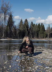 Mature woman playing with stick crouching at riverbank - JBYF00217