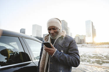 Lächelnder Mann, der im Winter sein Smartphone vor dem Auto benutzt - EYAF02513