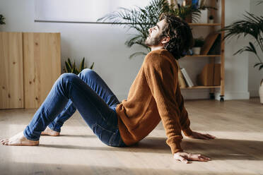 Man enjoying sunlight sitting with eyes closed on hardwood floor at home - EBBF07474