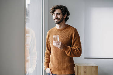 Happy young man with glass of water looking through window - EBBF07478