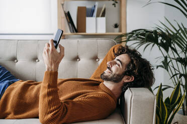 Glücklicher junger Mann, der sein Smartphone benutzt und sich auf dem Sofa zu Hause entspannt - EBBF07480