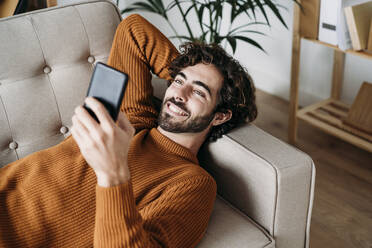 Glücklicher junger Mann mit Smartphone auf dem Sofa - EBBF07481