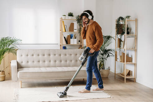 Happy young man wearing wireless headphones cleaning home with vacuum cleaner - EBBF07485