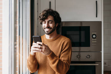 Happy young man using smart phone in kitchen at home - EBBF07489