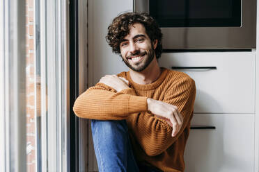 Smiling young man sitting by window in kitchen at home - EBBF07491