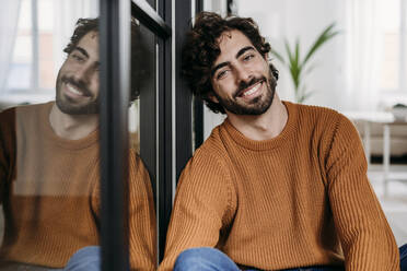 Happy young man leaning on glass door at home - EBBF07492