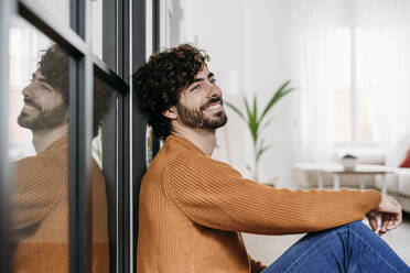 Thoughtful young man leaning on glass door at home - EBBF07493