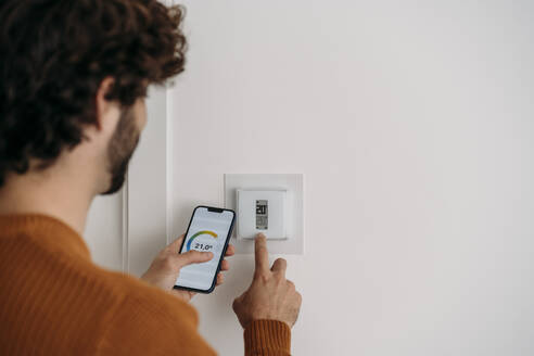 Junger Mann bedient Thermostat mit Smartphone zu Hause - EBBF07494