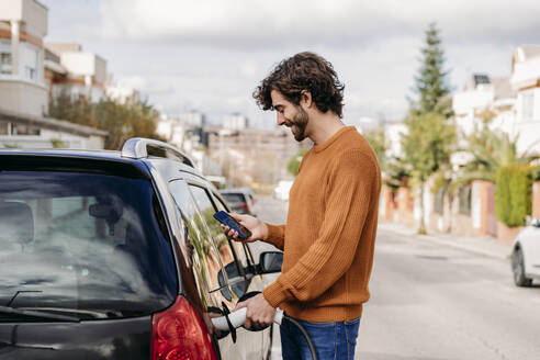 Junger Mann benutzt sein Smartphone und lädt sein Auto an einer Ladestation für Elektrofahrzeuge - EBBF07499