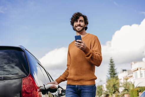 Glücklicher junger Mann mit Mobiltelefon, der sein Auto an einer Ladestation für Elektrofahrzeuge auflädt - EBBF07502