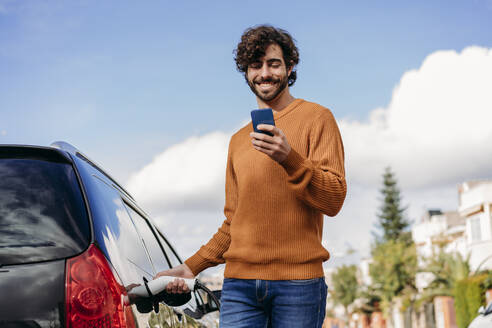 Happy man using smart phone and charging car at vehicle charging station - EBBF07503