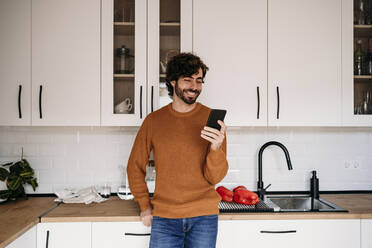 Happy man using smart phone leaning on kitchen counter at home - EBBF07511