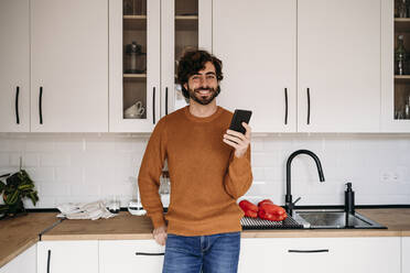 Happy man with smart phone leaning on kitchen counter at home - EBBF07512