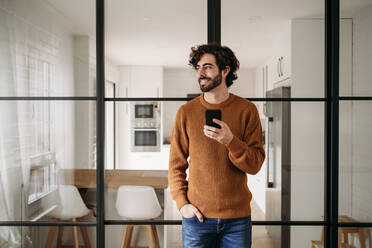 Happy young man with smart phone leaning on glass door at home - EBBF07515