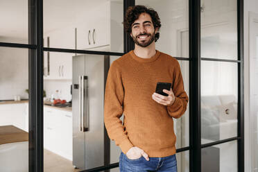 Smiling young man standing with hand in pocket and holding smart phone at home - EBBF07517