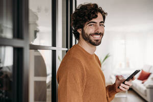 Happy man with smart phone standing by glass door at home - EBBF07518