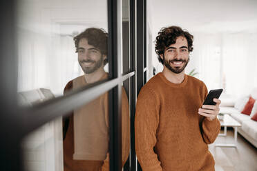 Smiling man with mobile phone standing by glass door at home - EBBF07520