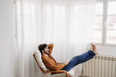 Glücklicher junger Mann entspannt sich auf einem Sessel im Schlafzimmer zu Hause - EBBF07522