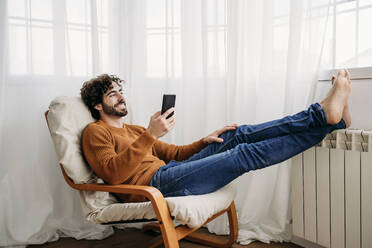 Glücklicher Mann mit Smartphone, der zu Hause auf einem Sessel sitzt - EBBF07525