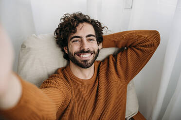 Happy man taking selfie on armchair at home - EBBF07526