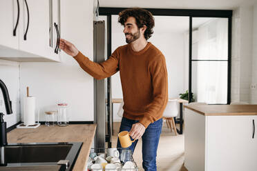 Happy young man with cup opening cabinet in kitchen at home - EBBF07545