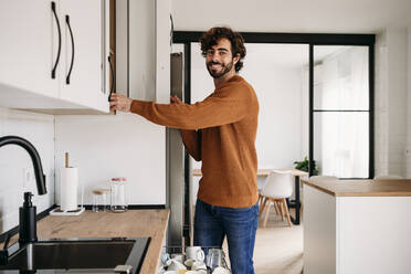 Happy man opening cabinet in kitchen at home - EBBF07546