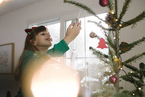 Lächelndes Mädchen hält eine Weihnachtskugel, die zu Hause am Weihnachtsbaum hängt - TYF00628