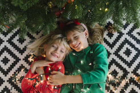 Fröhliche Schwestern mit beleuchteter Lichterkette, die in der Nähe des Weihnachtsbaums liegen - TYF00631