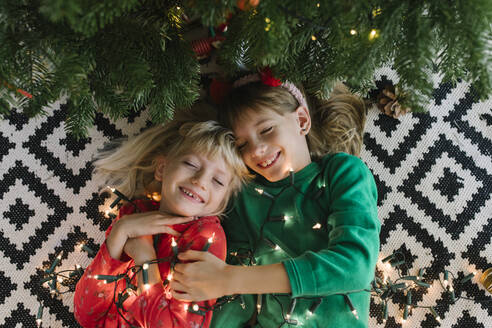 Happy sisters with illuminated string lights lying down near Christmas tree - TYF00631