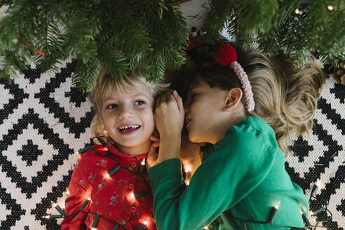 Mädchen flüstert seiner Schwester ins Ohr und legt sich mit einer Lichterkette in der Nähe des Weihnachtsbaums hin - TYF00632