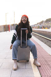 Glückliches Mädchen mit Strickmütze auf einem Koffer sitzend am Bahnsteig - JAQF01116