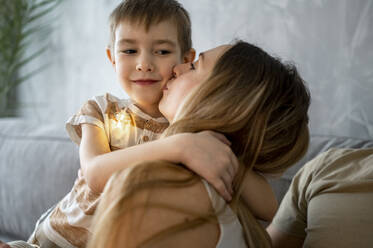 Mother kissing son sitting on sofa at home - ANAF00799