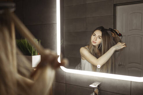 Frau lockt Haare mit einer Zange vor einem Spiegel zu Hause - ANAF00800