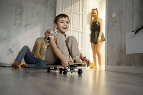 Vater spielt mit seinem Sohn, der auf einem Skateboard sitzt, während die Mutter im Hintergrund steht - ANAF00814