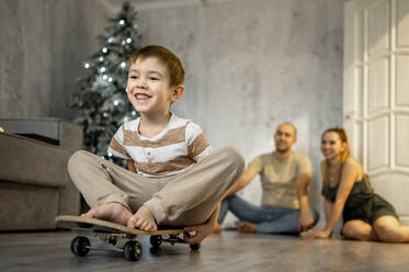 Happy father and mother playing with son sitting on skateboard at home - ANAF00815