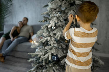 Junge beim Schmücken des Weihnachtsbaums mit Vater und Mutter auf dem Sofa sitzend im Hintergrund - ANAF00820
