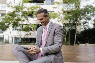 Lächelnder Geschäftsmann, der vor einem Gebäude eine Textnachricht auf seinem Smartphone verschickt - RFTF00324