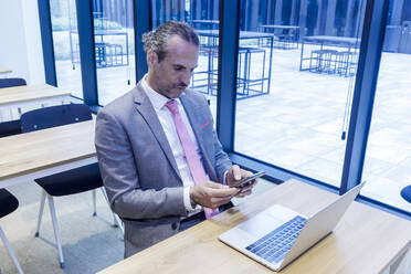 Mature businessman using smart phone by laptop at desk in office - RFTF00331