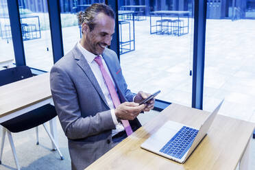 Glücklicher reifer Geschäftsmann, der sein Smartphone am Laptop am Schreibtisch im Büro benutzt - RFTF00333