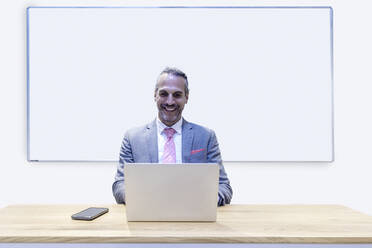 Lächelnder Geschäftsmann, der an einem Laptop vor einem Whiteboard im Büro sitzt - RFTF00335